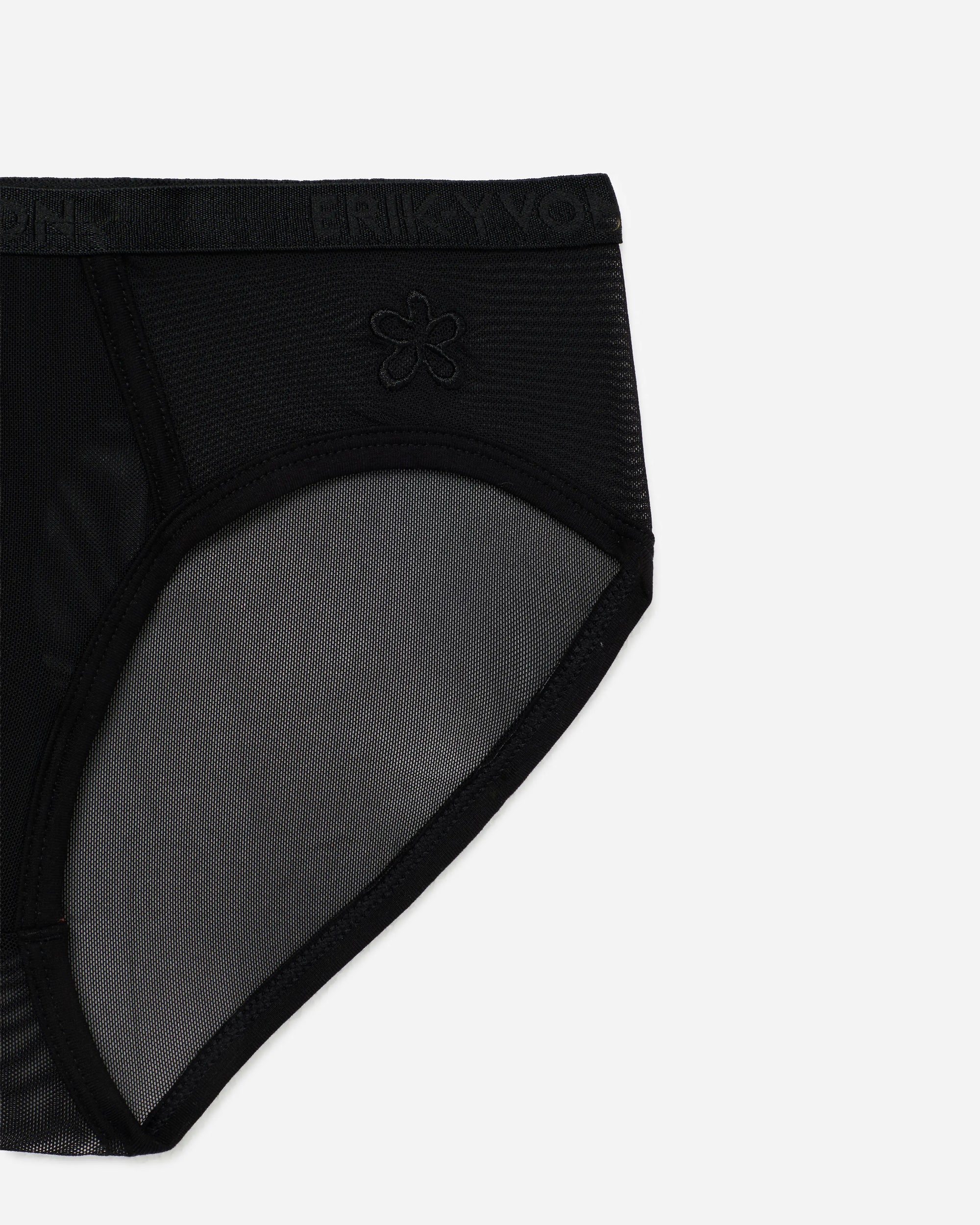 Close-up of a black sheer mesh and ribbed fabric brief, showing the inner leg panel and a small tonal embroidered flower logo on a light gray background.