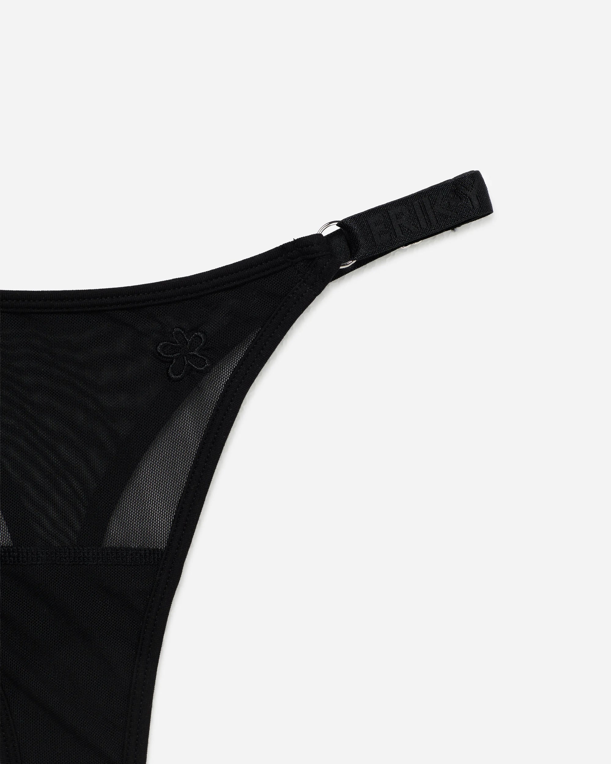 Close-up of a black sheer mesh thong with a wide adjustable strap and a small tonal embroidered flower detail, shown on a light gray background.